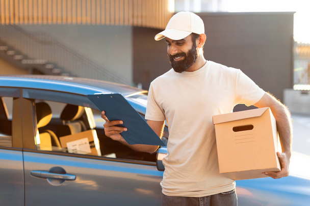 Delivery Offer. Happy Arab Deliveryman Holding Cardboard Parcel Box And Looking At Clipboard Standing Near Car Outdoors, Wearing Cap. Shot Of Courier Delivering Customers Orders - Photo, Image