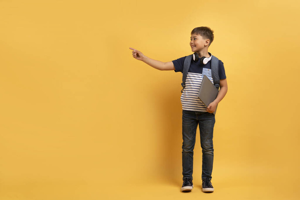 Cute cheerful happy kid wearing casual outfit, carrying backpack and books schoolboy with wireless headphones showing pointing at copy space, smiling, isolated on yellow background. Online education - Photo, Image