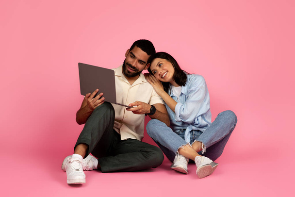 Loving beautiful young multiethnic couple using laptop computer on pink background, happy millennial man and woman sitting on floor, bonding and watching movie online, websurfing, scrolling together - Photo, Image