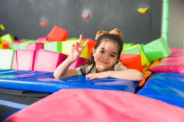 Beautiful happy kid smiling and making the peace sign while having fun playing in the trampoline area of the playroom - 写真・画像