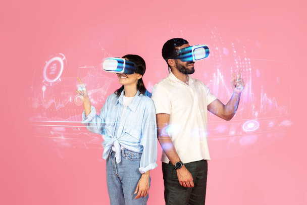 Счастливые возбужденные молодой человек и женщина, используя очки VR, трогательные голограммы игровой интерфейс, пара наслаждаться игрой в виртуальной реальности, розовый фон, студия. Веселье, развлечения с технологиями, коллаж - Фото, изображение