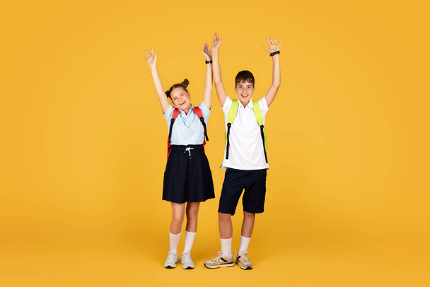 Iloinen valkoihoinen teini tyttö ja poika oppilas nostaa kätensä ylös, juhlii voittoa ja menestystä, eristetty oranssi studio tausta, täyspitkä. Opiskele yhdessä, kouluopetus, mainos ja tarjous - Valokuva, kuva