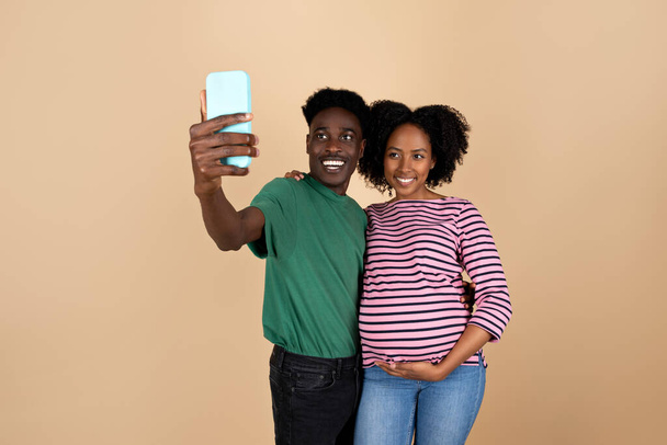 Vrolijke jonge zwarte man knuffelende dame met grote buik, selfie maken op smartphone, geïsoleerd op beige achtergrond, studio. Ouders en familie, relatie, app voor sociale media, zwangerschap - Foto, afbeelding