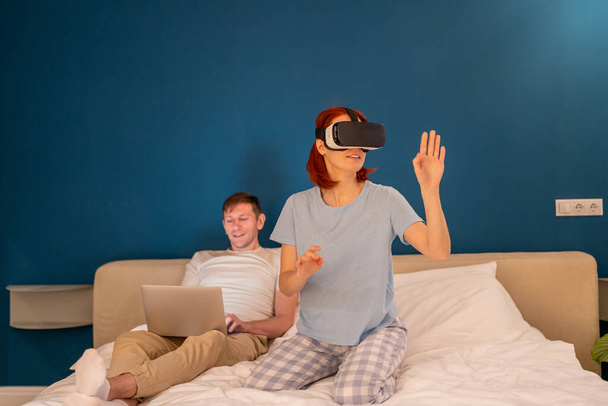 Mujer interesada en gafas de realidad virtual se sienta en la cama con el marido maneja gafas vr desde el ordenador portátil. Joven hembra en auriculares 3D explorando el mundo metaverso ciberespacio tocando cosas irreales jugando juego - Foto, imagen