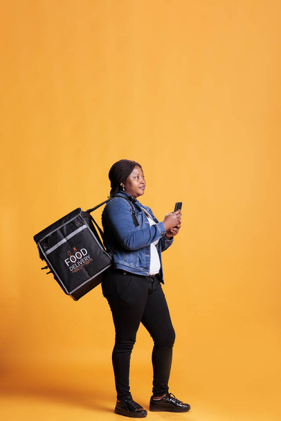 Afroamerykanka dostawy trzyma telefon patrząc na adreess klienta na aplikacji fast food przed rozpoczęciem dostawy zamówienia na wynos. Kurier pizzerii niosący plecak termiczny na wynos w studio - Zdjęcie, obraz