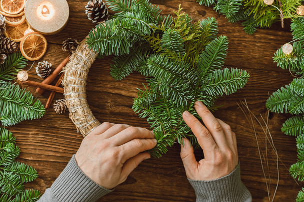 Männerhände basteln aus naturbelassenen Fichtenzweigen und verschiedenen Dekorationen einen Weihnachtskranz.  - Foto, Bild