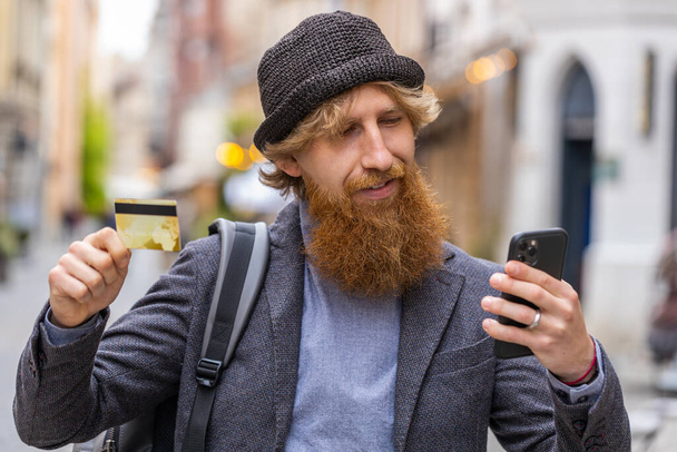 Молодий бородатий чоловік використовує смартфон кредитних карток при переказі грошей, покупках в Інтернеті, замовленні доставки їжі, бронюванні номеру готелю. Рудий хлопець, що йде в міському місті вулиці на відкритому повітрі
 - Фото, зображення