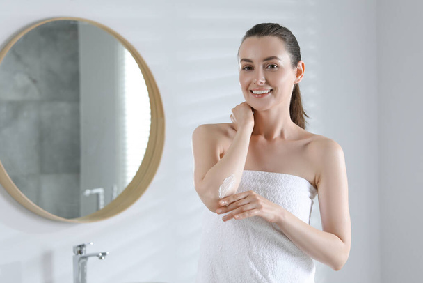 Happy woman applying body cream onto elbow in bathroom, space for text - Photo, image
