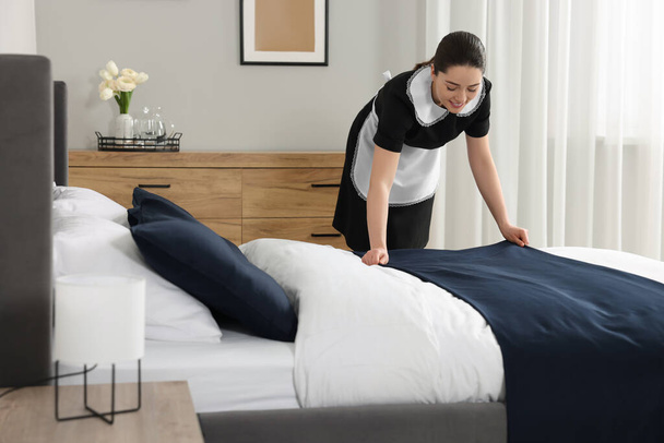 Young chambermaid making bed in hotel room - 写真・画像
