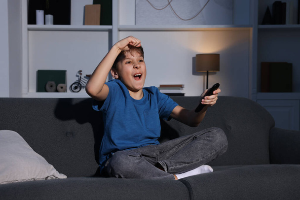 Érzelmes fiú néz TV-t és tartja távirányító kanapén otthon - Fotó, kép