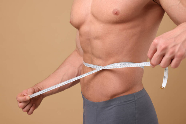Athletic man measuring waist with tape on brown background, closeup. Weight loss concept - Foto, Imagen