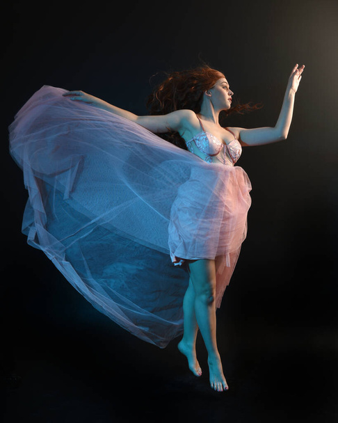 Ολόσωμο πορτραίτο όμορφης μελαχρινής χορεύτριας, με αιθέριο φόρεμα, χορό με ρέον ύφασμα. απομονωμένο σε σκοτεινό φόντο στούντιο με κινηματογραφικό πολύχρωμο φωτισμό. - Φωτογραφία, εικόνα