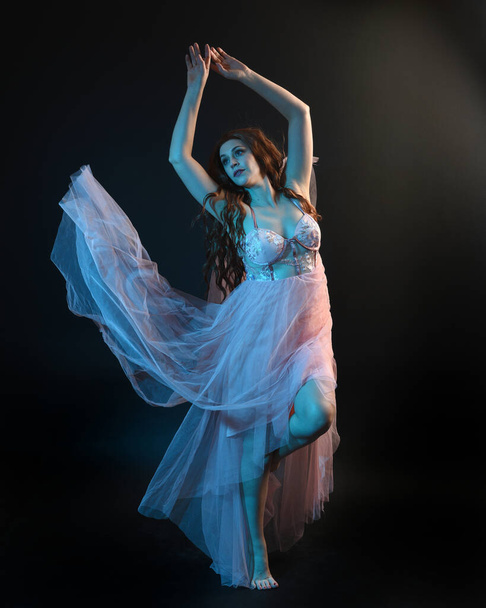 Ganzkörperporträt der schönen brünetten Tänzerin, die ätherische Kleider trägt und mit fließendem Stoff tanzt. isoliert auf dunklem Studiohintergrund mit cineastischer farbenfroher Beleuchtung. - Foto, Bild