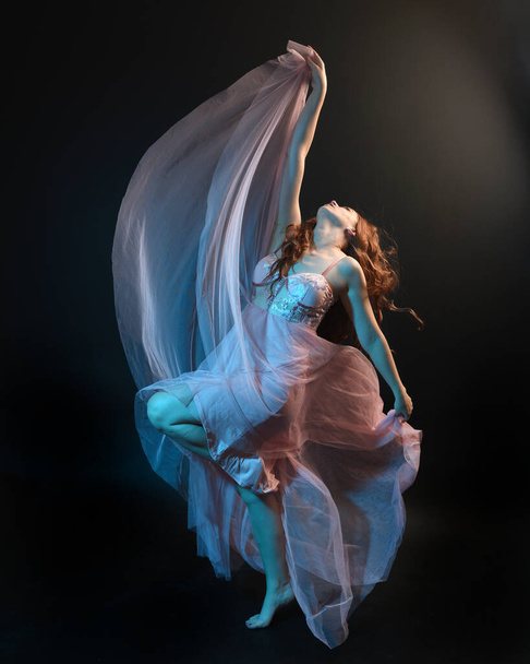 美しいブルネットの女性ダンサーの完全な長さの肖像画は,イーサリアルガウンを着て,流れる生地で踊ります. 映画のカラフルな照明とダークスタジオの背景に隔離された. - 写真・画像