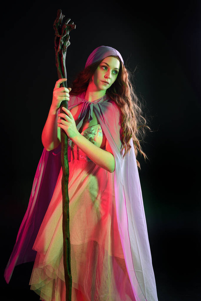 Retrato de larga duración de una hermosa morena vestida con un vestido con capa de fantasía púrpura sosteniendo un mago de madera, aislado en el fondo del estudio oscuro con iluminación colorida cinematográfica. - Foto, imagen