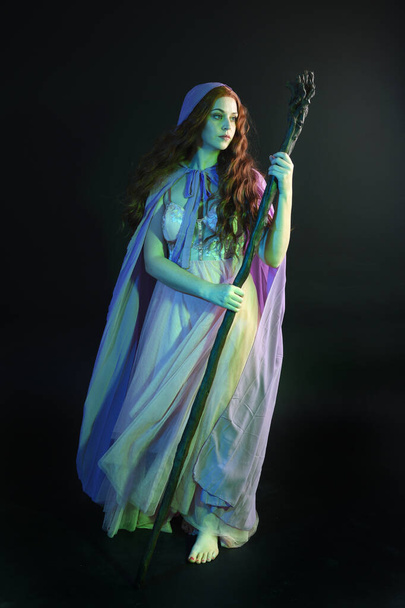 Full length portrait of beautiful brunette woman wearing  a gown with purple fantasy cloak holding a wooden wizard staff, isolated on dark studio background with cinematic colourful lighting. - Photo, Image