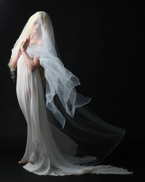 Полноразмерный портрет красивой блондинки в белом платье с текущей призрачной завуалированной тканью, изолированной на фоне дарской студии. - Фото, изображение