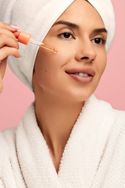Positive young female in bathrobe and towel on head applying serum oil on skin on pink background - Фото, зображення