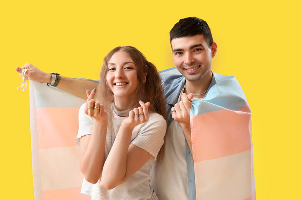 Νεαρό ζευγάρι με τρανσέξουαλ σημαία κάνοντας σχήμα καρδιάς με δάχτυλα σε κίτρινο φόντο - Φωτογραφία, εικόνα