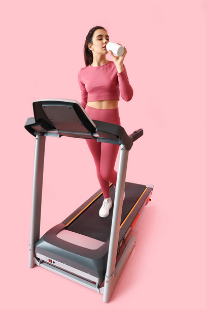 Sporty young woman drinking water on treadmill against pink background - Фото, зображення