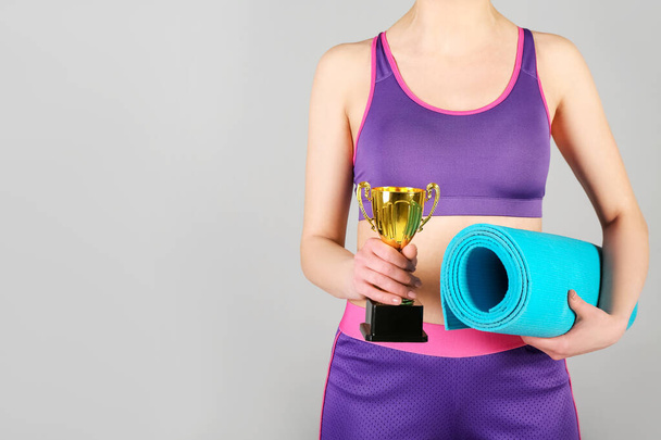 Αθλητική νεαρή γυναίκα με fitness mat και χρυσό κύπελλο σε γκρι φόντο - Φωτογραφία, εικόνα