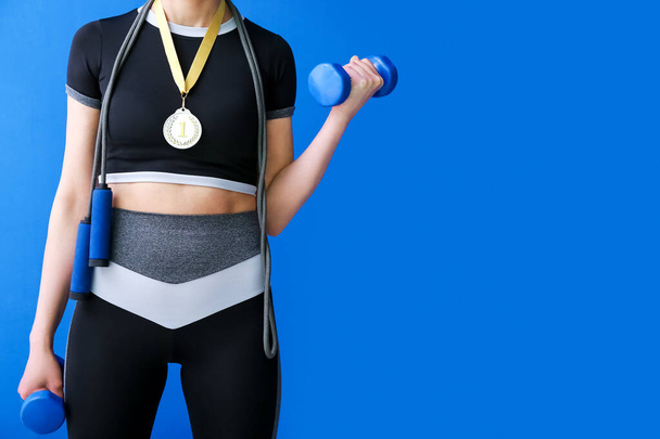 Sporty young woman with first place medal, skipping rope and dumbbells on blue background - Photo, Image