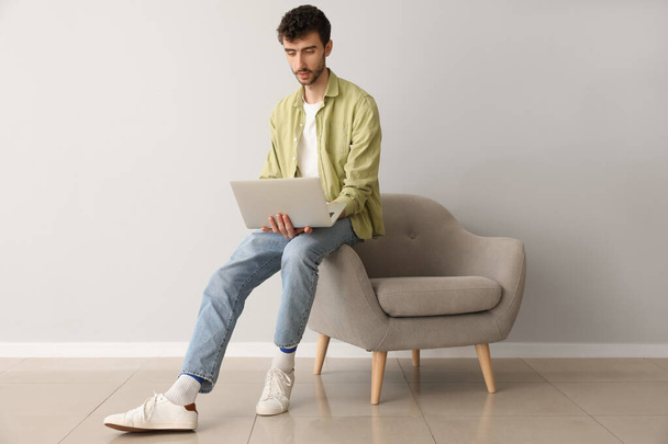 Молодой человек работает с ноутбуком на кресле возле светлой стены - Фото, изображение