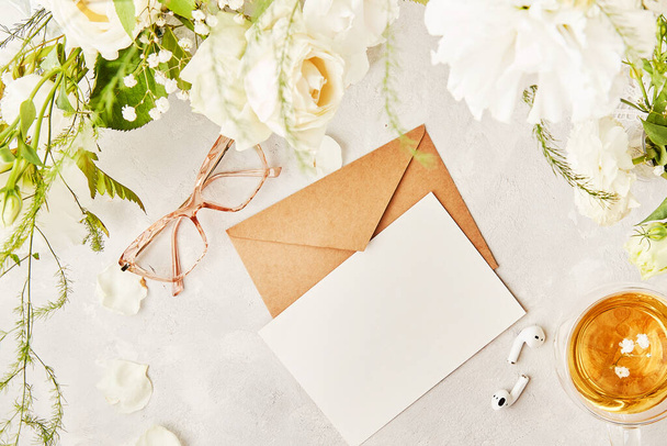 O apartamento feminino põe-se com a elaboração de envelope de papel pastel de cumprimentos, convite, texto entre flores brancas. Óculos, xícara de chá e fones de ouvido vista superior. - Foto, Imagem