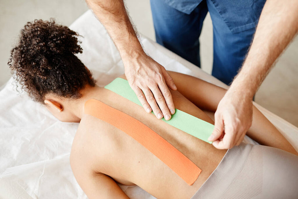 Top weergave van mannelijke revalidatie specialist zetten fysio tape op de rug van jonge vrouw, kopieerruimte - Foto, afbeelding