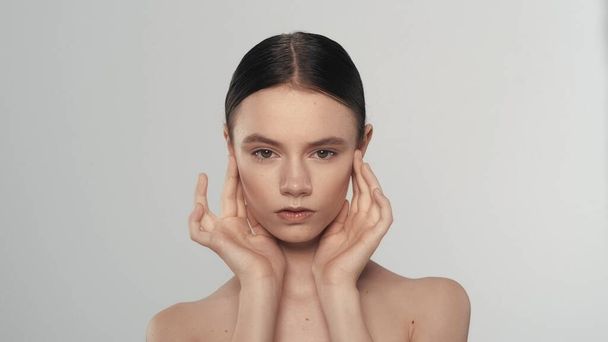 Portrait of a beautiful female model with ponytail. Close up shot of a woman with nude makeup and hands up, touching her face soft skin, looking at the camera. Beauty advertisement concept. - 写真・画像