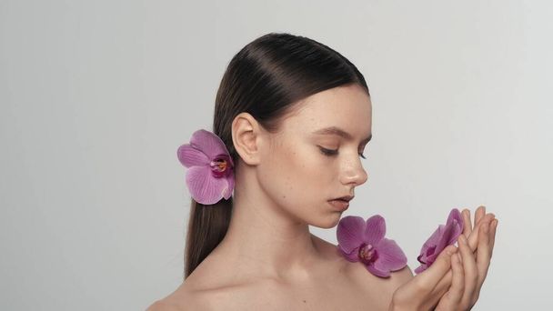 Portrait of a appealing brunette female model. Close up shot of a woman with ponytail holding an orchid flower in her hand and looking at it. Organic skincare advertisement concept. - 写真・画像