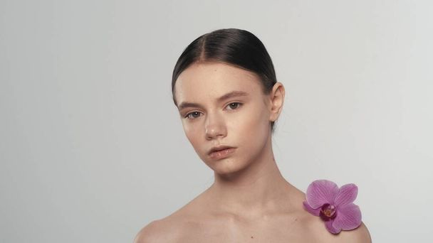 Portrait of appealing brunette female model. Close up shot of a woman with ponytail holding an orchid flower on her shoulder looking at the camera. Organic skincare advertisement concept. - 写真・画像