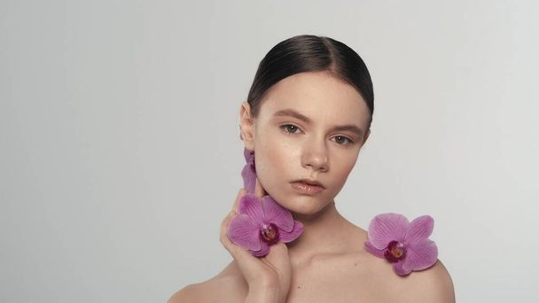 Portrait of caucasian female model. Close up shot of a woman with ponytail holding an orchid flower in her hand and looking at the camera. Organic skincare advertisement concept. - 写真・画像