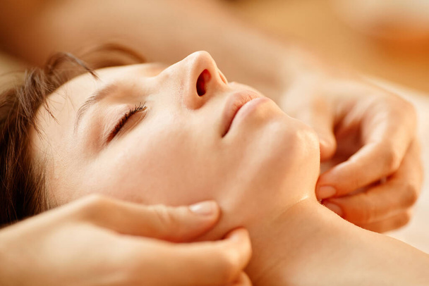 Ciepłe stonowane zbliżenie młodej kobiety korzystającej z masażu twarzy w centrum SPA z zamkniętymi oczami - Zdjęcie, obraz