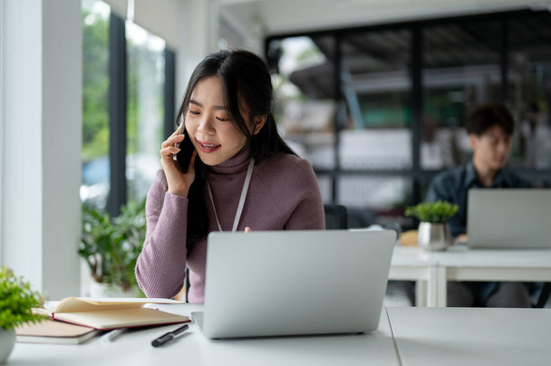 Egy gyönyörű és profi ázsiai üzletasszony vagy női értékesítési asszisztens beszél telefonon egy ügyféllel, miközben az íróasztalánál ül az irodában.. - Fotó, kép
