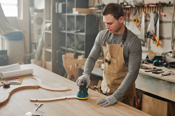 Портрет молодого плотника, шлифующего деревянную мебель ручной работы в туманной мастерской, копировальное пространство - Фото, изображение