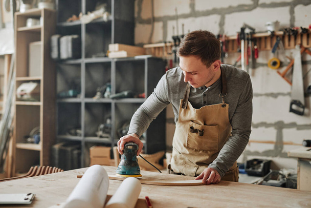 Retrato cálido tonificado de carpintero joven lijando madera y construyendo muebles hechos a mano en taller, espacio para copiar - Foto, Imagen
