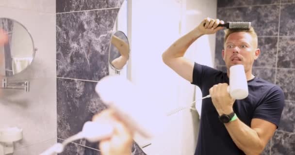 Giovane uomo pettinando davanti allo specchio e soffiando le guance con asciugacapelli film 4k. Scelta di asciugacapelli dal concetto di potenza - Filmati, video