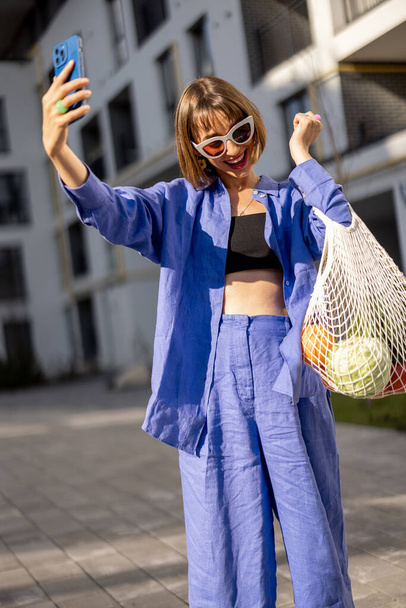 Mujer feliz habla por teléfono en línea mientras camina a casa con una bolsa de malla llena de frutas y verduras frescas en el patio interior del edificio de apartamentos - Foto, imagen