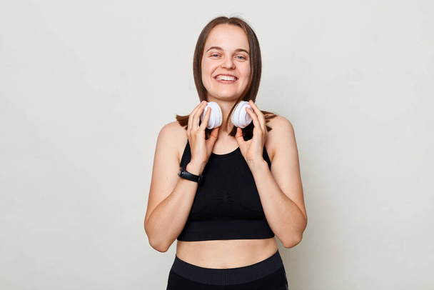 Smiling positive cheerful slim woman with headphones wearing sportswear posing against gray background lookign at camera with positive expression. - Valokuva, kuva