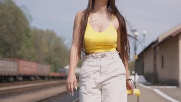 Улыбающаяся туристка в ярко-желтой шляпе и солнечных очках покоряет железнодорожную платформу с чемоданом, готовая к новым впечатлениям. Молодая женщина отправляется в отпуск. Высокий - Кадры, видео
