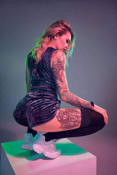 Beautiful young blonde tattoed girl in blue velour booty shorts and t-shirt, making squats on top of big cube, showing off her amazing booty - Zdjęcie, obraz
