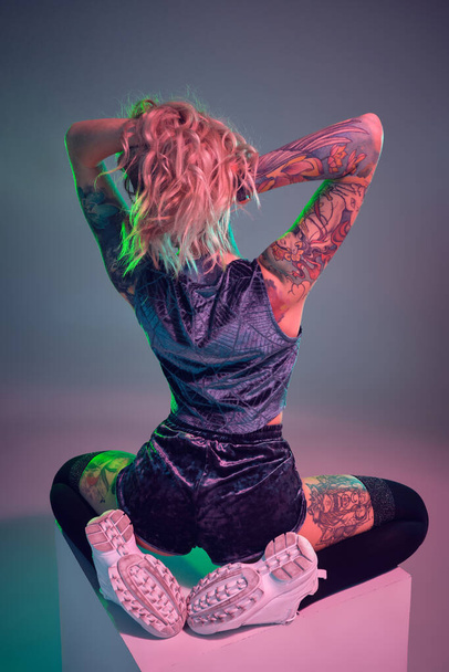 Beautiful young blonde tattoed girl in blue velour booty shorts and t-shirt, she sits on top of big cube, showing off her amazing booty from behind - Foto, Imagen