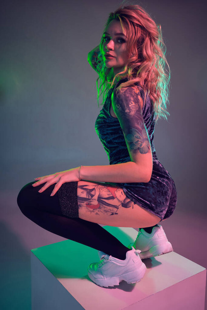 Beautiful young blonde tattoed girl in blue velour booty shorts and t-shirt, making squats on top of big cube, showing off her amazing booty - Foto, afbeelding