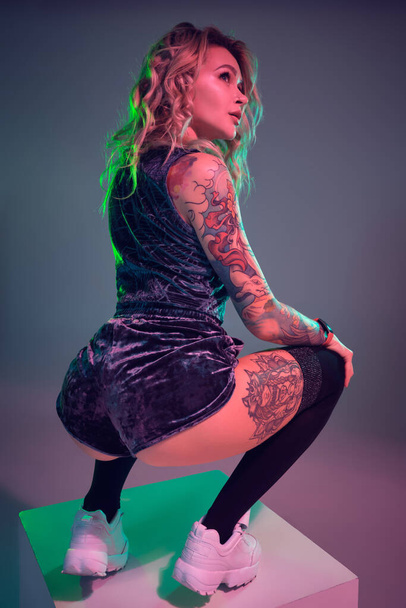 Beautiful young blonde tattoed girl in blue velour booty shorts and t-shirt, making squats on top of big cube, showing off her amazing booty - Valokuva, kuva
