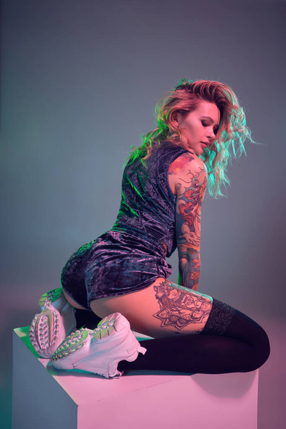Beautiful young blonde tattoed girl in blue velour booty shorts and t-shirt, she sits on top of big cube, showing off her amazing booty from behind - Foto, immagini