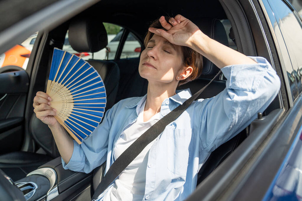 Exhausted tired middle aged woman drives car waves blue fan suffers from stuffiness stands in urban traffic jam in summer hot weather. Overheating, high temperature in car with broken air conditioner. - Photo, Image