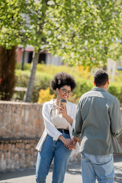 cheerful african american woman holding coffee to go and hand of boyfriend on urban street - Zdjęcie, obraz
