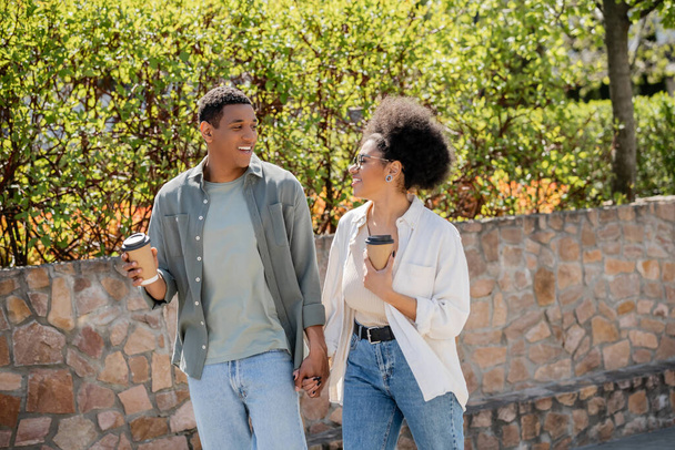 smiling african american couple holding coffee to go while talking and walking on urban street - Photo, Image
