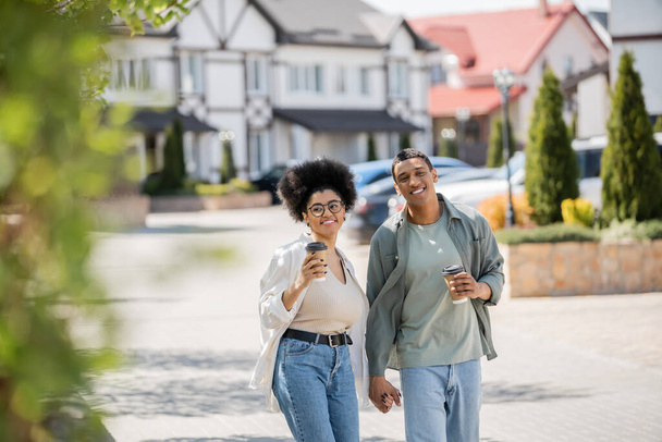 donna afro-americana sorridente con caffè per andare a passeggiare con il fidanzato vicino alle case sulla strada urbana - Foto, immagini